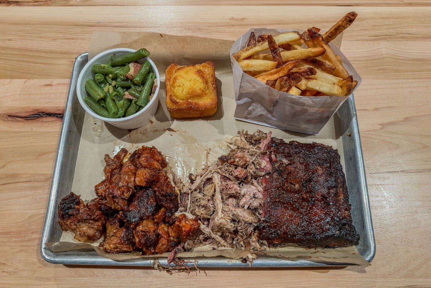 Essens-Tablett in einem Restaurant in Waterloo, Ontario. Das Menü ist sehr fleischlastig mit Burnt Ends (marinierte Schweinebrust-Würfel), Pulled Porc und St. Louis Style Ribs. Dazu Pommes frites in Papier, grüne Bohnen, ein Stück süsses Maisbrot und Barbecue-Sauce auf braunem Papier, serviert auf einem Holztisch.
