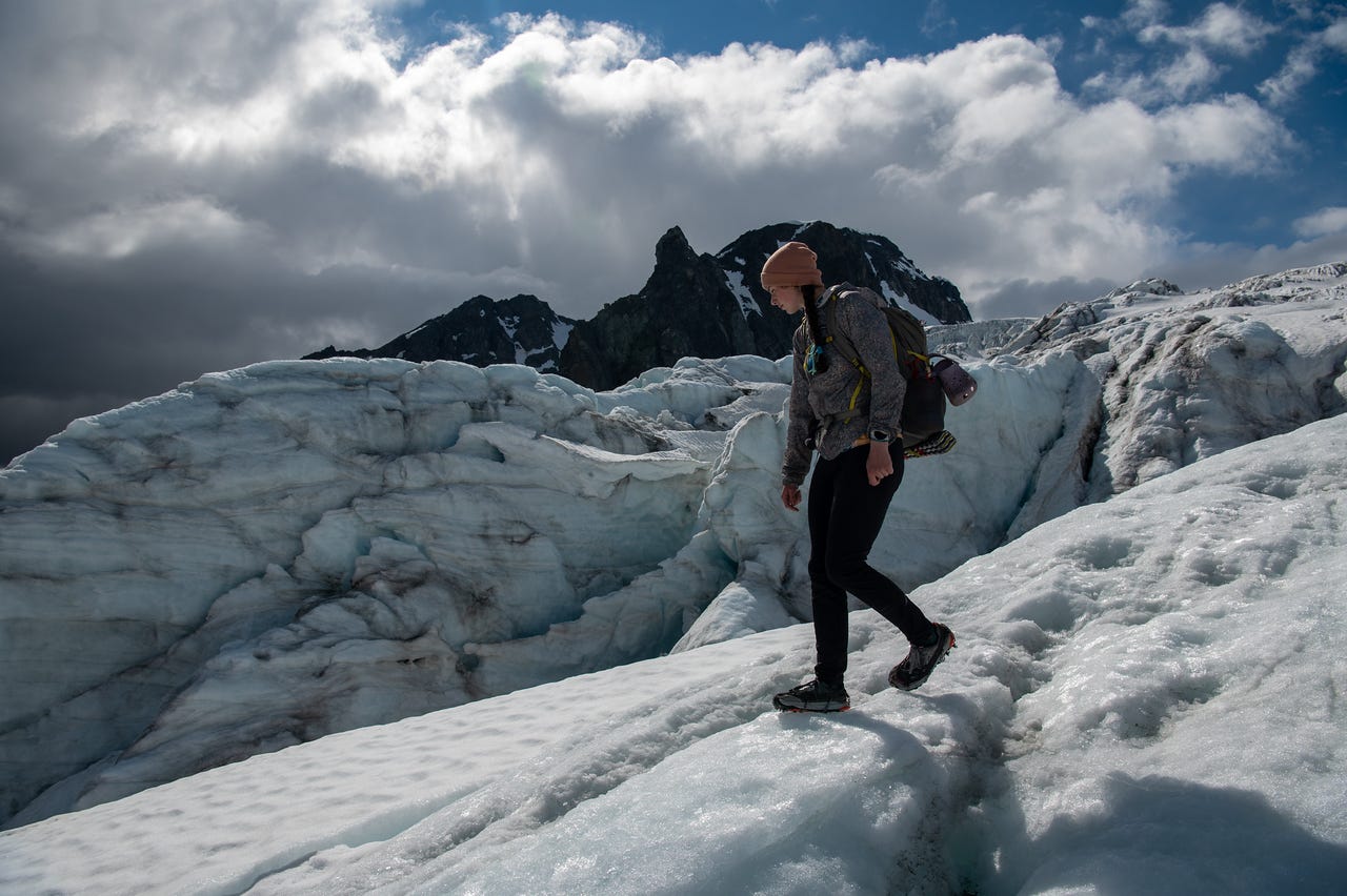 A girl in a peach beanie and grey hoody steps carefully down sunlit glacier ice in microspikes, a foam pad and water bottle strapped to her pack, her shadowed profile concentrated on the slope. Behind her, walls of fractured blue ice rise toward a dark rock peak, with brilliant white cloud and a patch of blue sky above and a nearly black storm cloud at left.