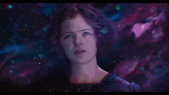 A red-haired woman floats against a swirling purple-pink cosmic background, her face illuminated by shimmering lights as she gazes forward and blinks slowly.