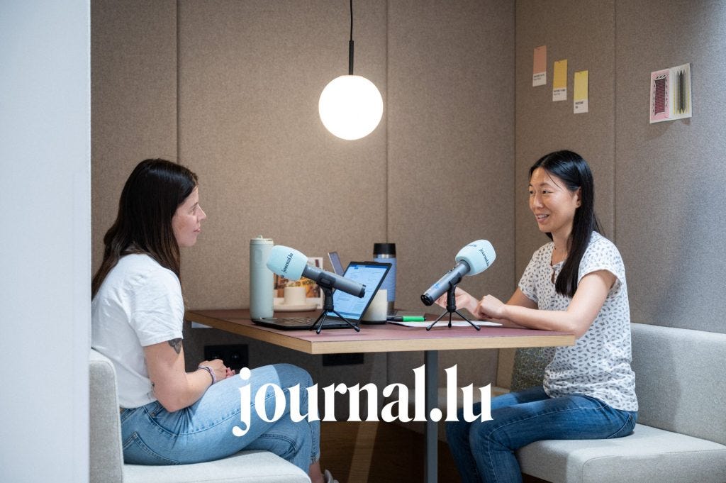 Two people sit in a small booth recording a podcast, speaking into microphones across a table with laptops, notebooks, and water bottles under a hanging round light.