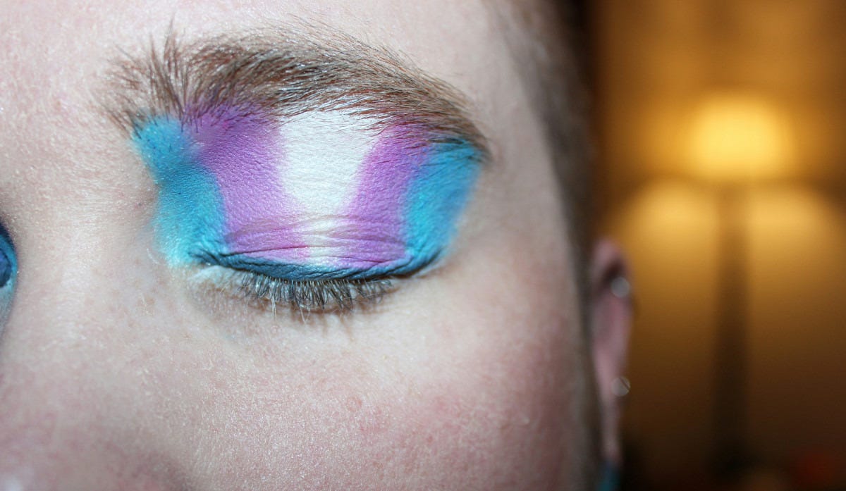 Closeup of a person’s face with an eyelid painted in the colors of the pro-trans movement.