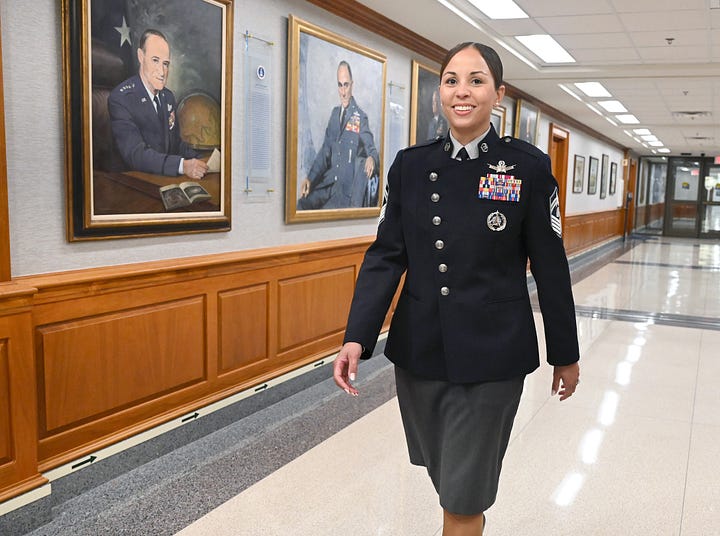 A smiling U.S. Space Force service member walks down a bright, polished hallway lined with wood paneling and framed portraits of military leaders. She wears a dark-blue dress uniform with silver buttons, senior enlisted chevrons, a chest full of ribbons, and a Space Force insignia, paired with a gray skirt. Fluorescent ceiling lights and glossy tile floors lead toward glass doors at the end of the corridor.