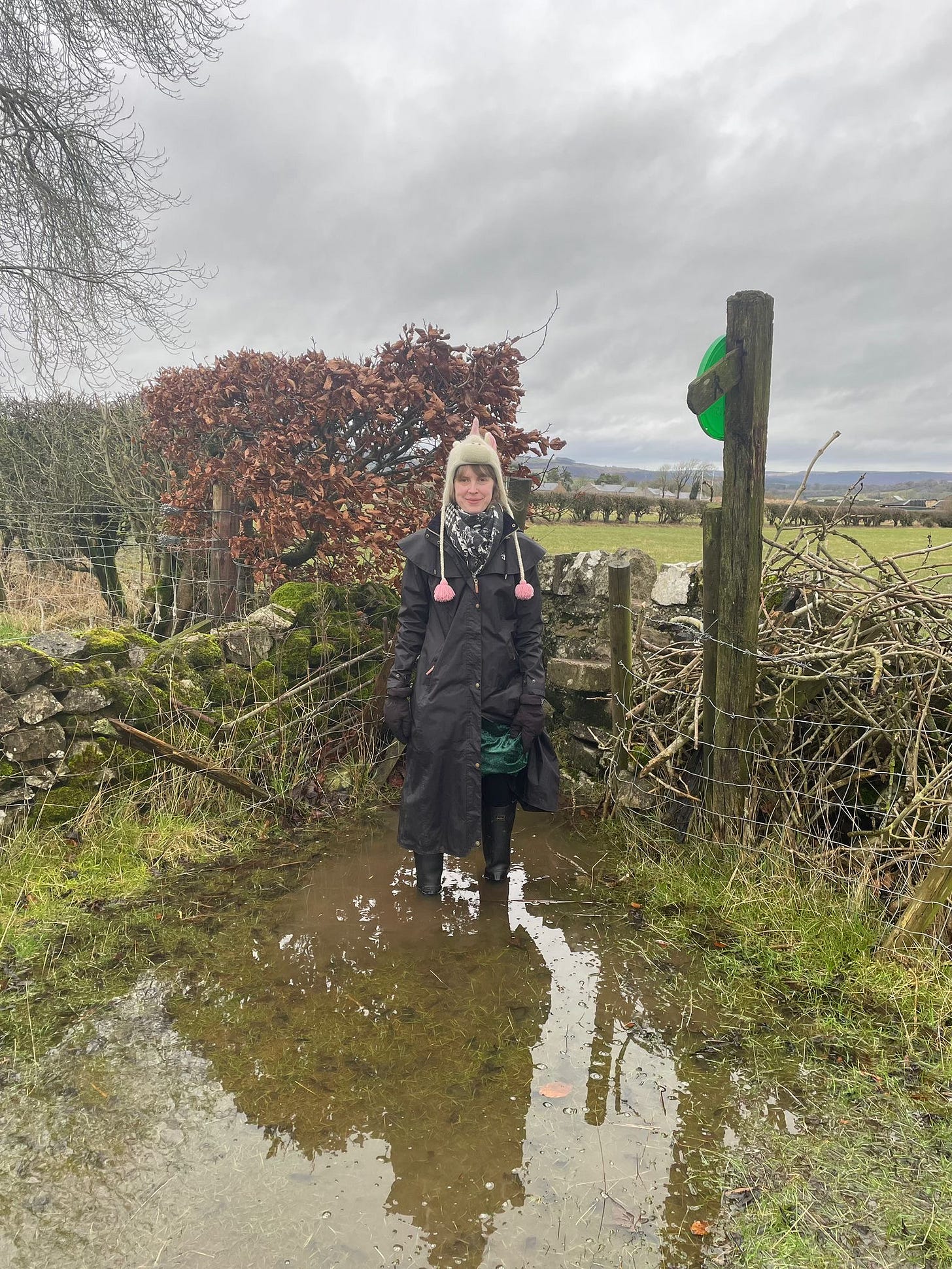 Photograph of the writer standing in a puddle, whilst wearing a long, black raincoat and black wellies. They are also wearing a wooly unicorn hat and smiling for the camera. It is a real smile. They like puddles.