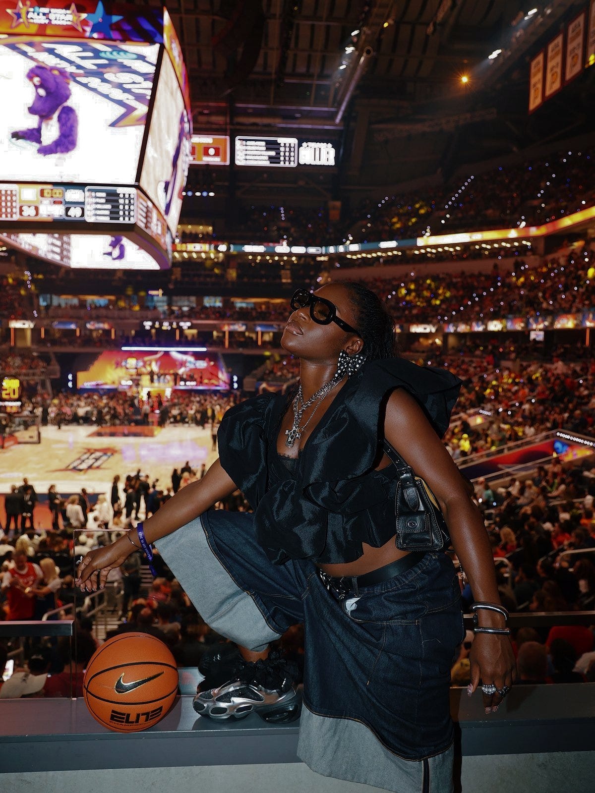 WNBA fan in an oversized fashion-forward outfit posing courtside inside a packed basketball arena WNBA fan in an oversized fashion-forward outfit posing courtside inside a packed basketball arena