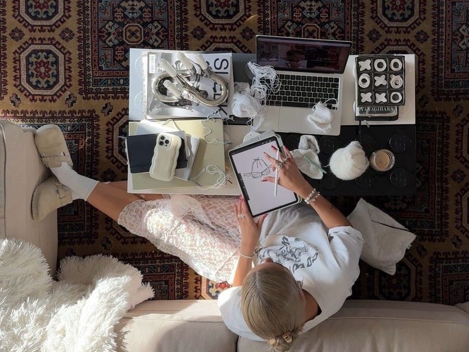 This may contain: a woman is sitting on the couch looking down at her laptop and other items that are scattered around her