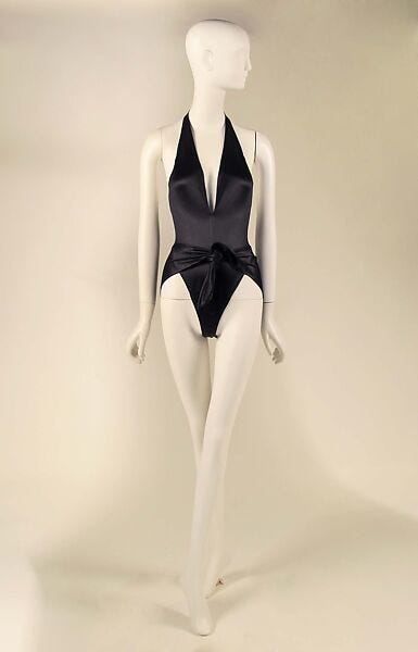 A mannequin wears a black swimsuit designed by Halston with a plunging neckline and high-cut leg openings that ties at the front waist.