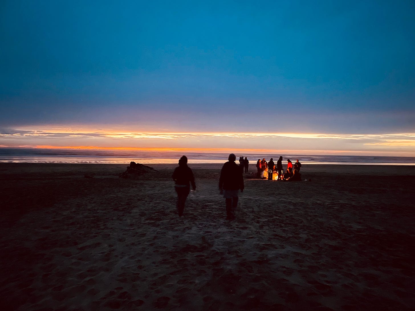 Photo of people in the distance and mostly in silhouette gathered around a bonfire on the beach at sunset with the ocean just behind them. The sky is orange with some clouds fading to a deep blue. Two people in the foreground walk away from the camera toward the fire. 