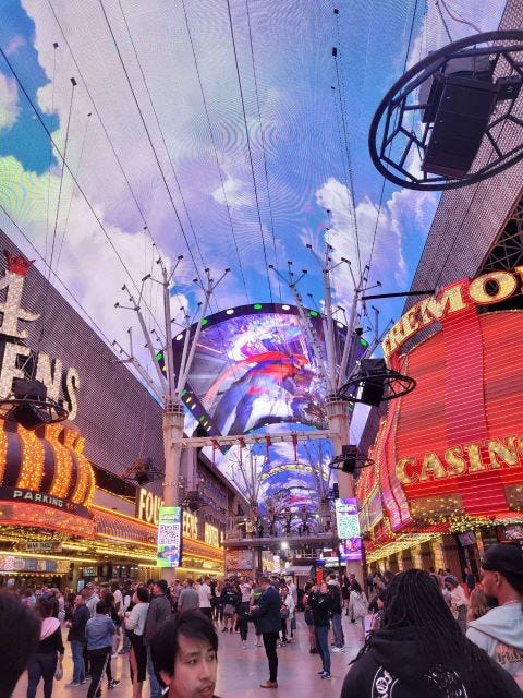 Foto da Frémont Street, com muita gente circulando no piso, cassinos cheios de neon dos lados e o teto é arredondado com a projeção de um céu com nuvens. Tudo é muito colorido.