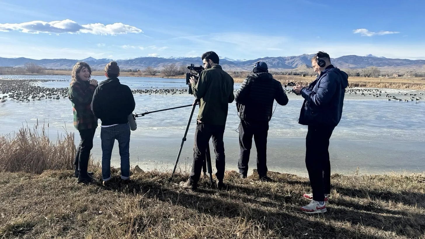 Megan, Andrea, plus Cinematographer, Sound Engineer, and Director, shooting at a lake in Longmont, Colorado. Geese on the water. Mountains in the background. Smiles, everyone. Megan, Andrea, plus Cinematographer, Sound Engineer, and Director, shooting at a lake in Longmont, Colorado. Geese on the water. Mountains in the background. Smiles, everyone.