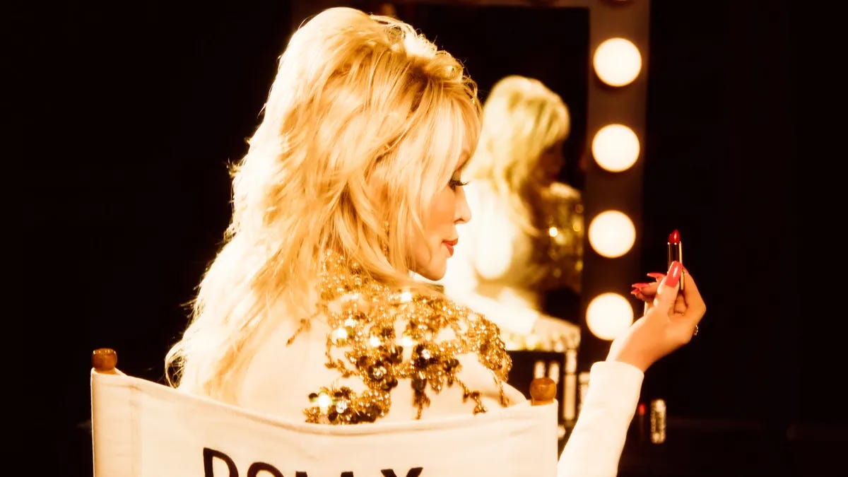 A person, Dolly Parton, is seen from behind holding a lipstick and sitting in chair that reads "Dolly" across the back. A person, Dolly Parton, is seen from behind holding a lipstick and sitting in chair that reads "Dolly" across the back.