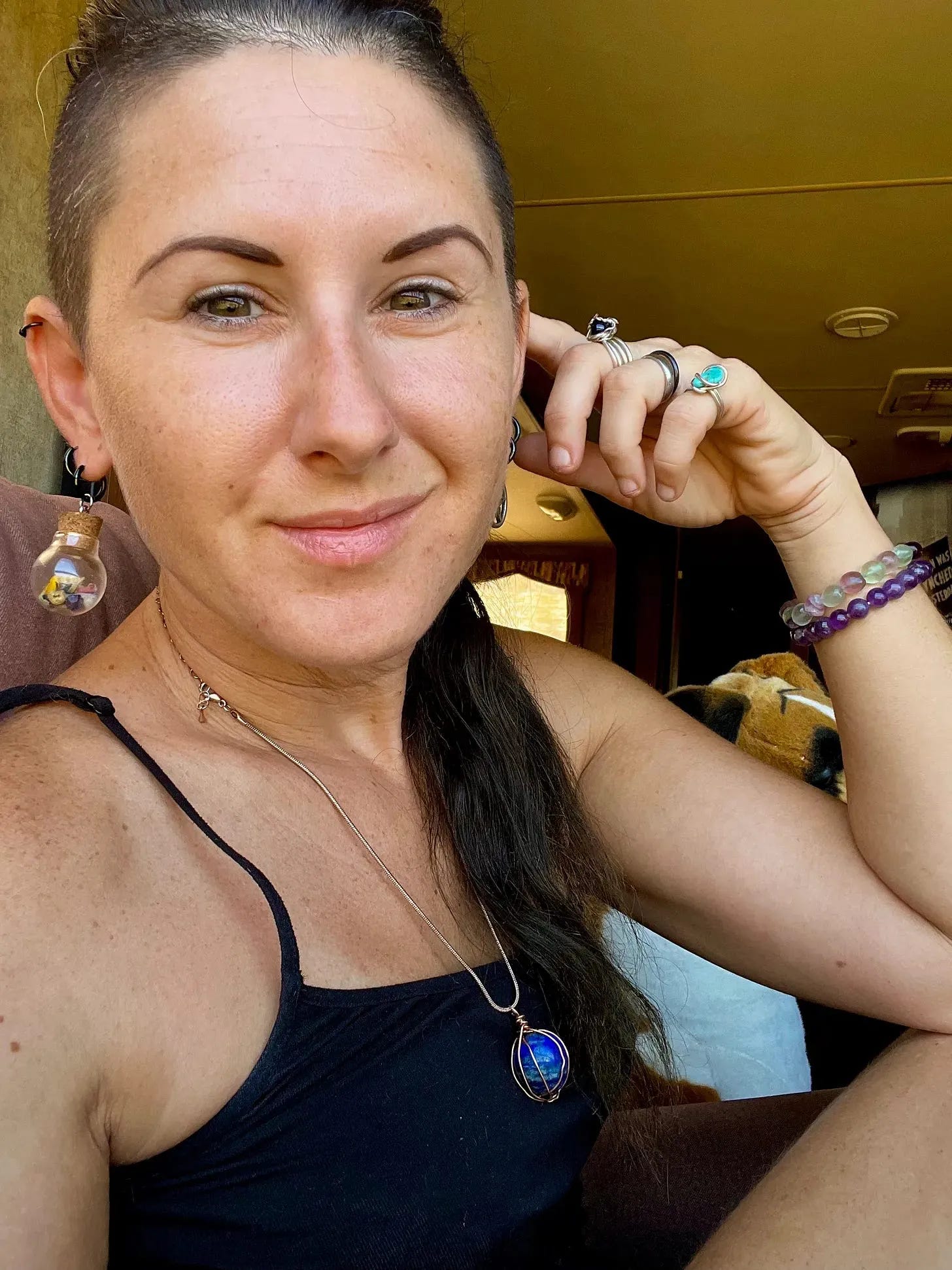 Lyric, posing in their RV. They are wearing a black spaghetti strap shirt, blue lapis necklace, two stone bracelets and multiple rings with various stones. 