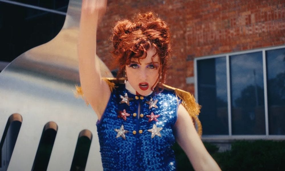 Chappell Roan, a white woman singer/songwriter with red curly hair. She's wearing red lipstick and mascara, and a sparkly blue majorette/cheerleader outfit with stars and gold epaulettes. She's looking at the camera and singing, in the middle of throwing a hand up in a dance. Chappell Roan, a white woman singer/songwriter with red curly hair. She's wearing red lipstick and mascara, and a sparkly blue majorette/cheerleader outfit with stars and gold epaulettes. She's looking at the camera and singing, in the middle of throwing a hand up in a dance.