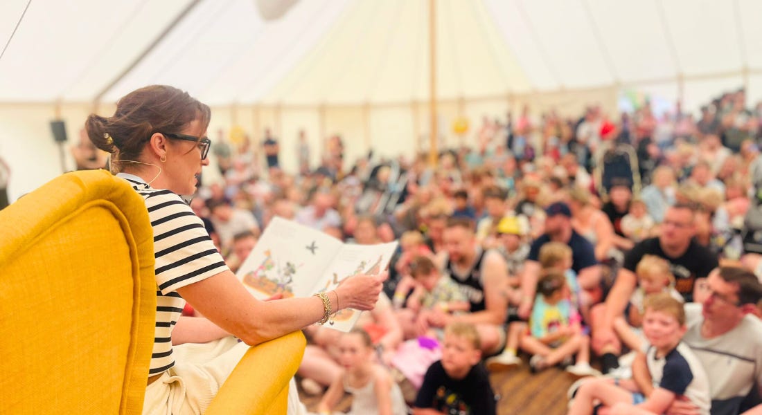 A woman with brown hair and a striped top sat in a yellow chair reading a book, with a crowd of people in front of her A woman with brown hair and a striped top sat in a yellow chair reading a book, with a crowd of people in front of her