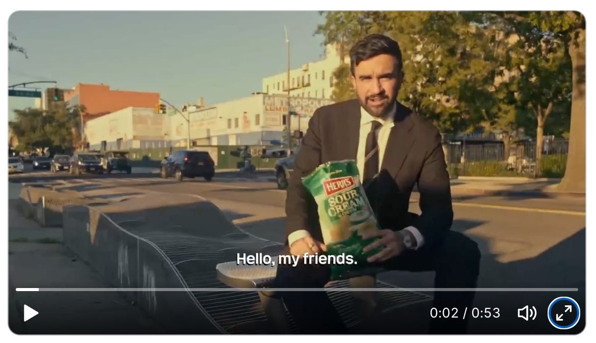 A screen grab of a video showing Zohran Mamdani, wearing a suit and casually seated on a sidewalk, holding a bag of Herr's sour cream and onion potato chips and speaking with his mouth full. The subtitle reads, "Hello, my friends."