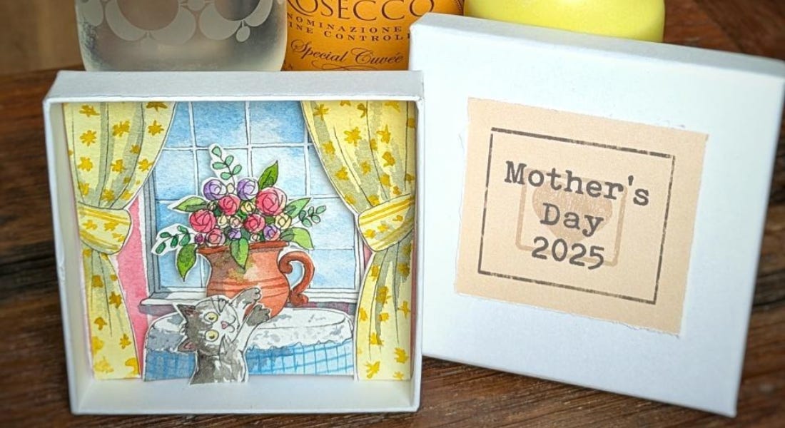 A diorama of a cat next to a vase of flowers and an art print that says 'mother's day 2025' stood up on a table