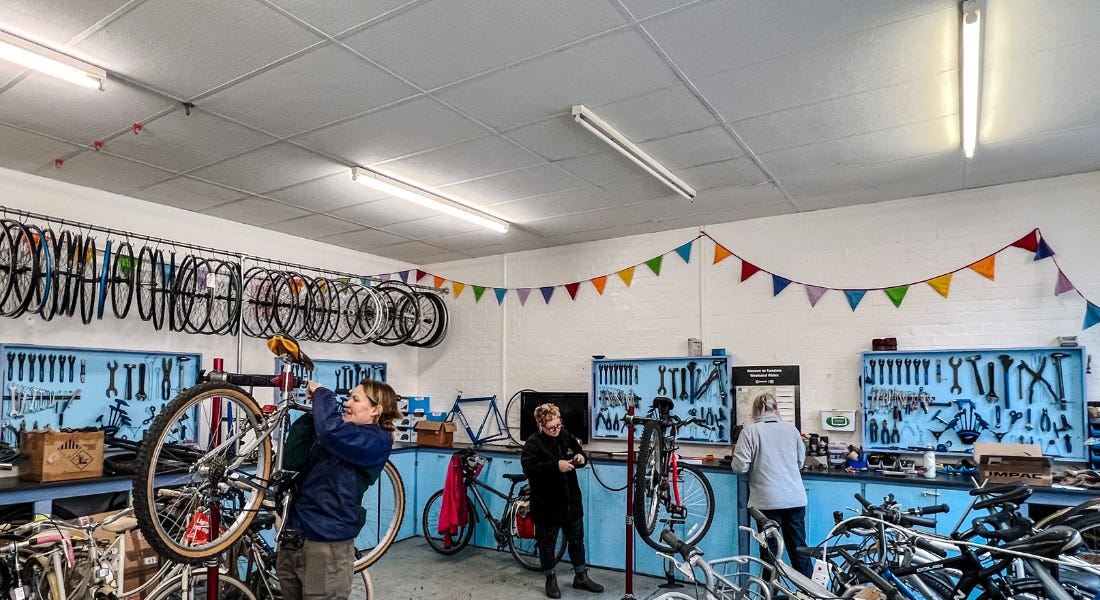 A bike workshop with bikes, tyres, and blue cabinets, and three women working on bikes within it A bike workshop with bikes, tyres, and blue cabinets, and three women working on bikes within it