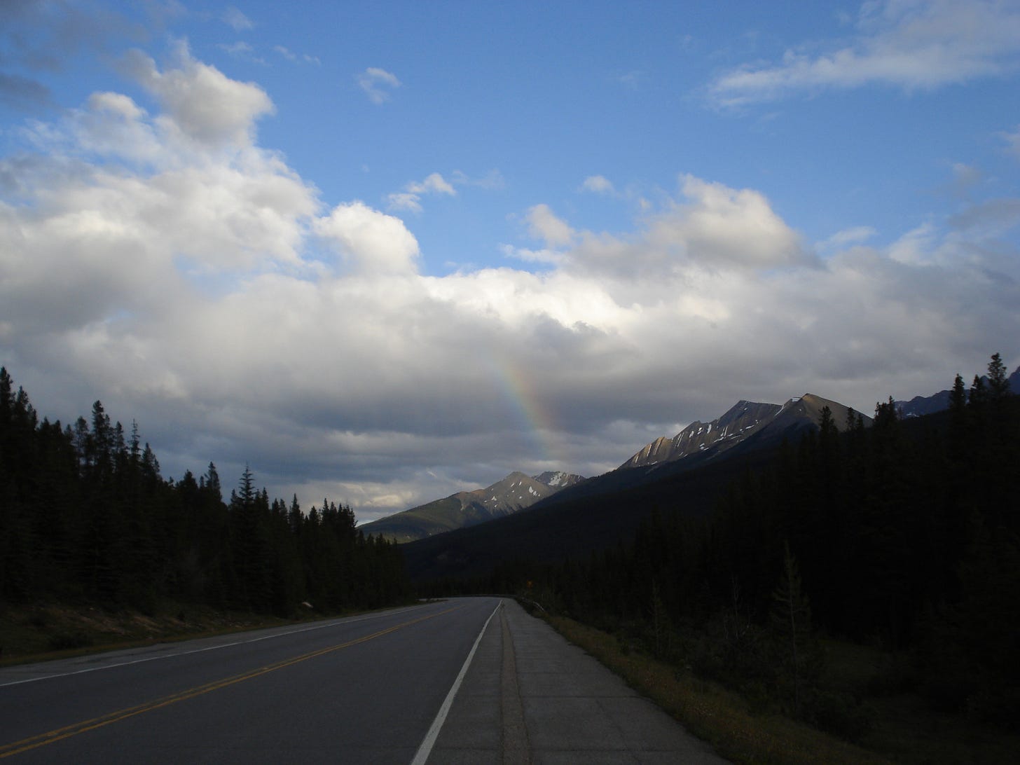 a rainbow peaks out from the clouds mixed with blue skies above a large mountain peak surrounded by trees