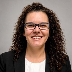 Retrato de uma mulher de cabelos cacheados escuros e compridos, usando óculos de armação azul, blazer preto e blusa branca. Ela sorri para a câmera diante de um fundo cinza neutro.