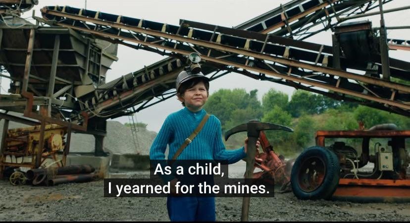 A still from the Minecraft movie. A Caucasian child between the ages of 10-12 wears a mining hat, a ribbed teal-colored long-sleeved sweater and holds a pickaxe in front of a mining site and giant cranes. Burned-in subtitles say “As a child, I yearned for the mines