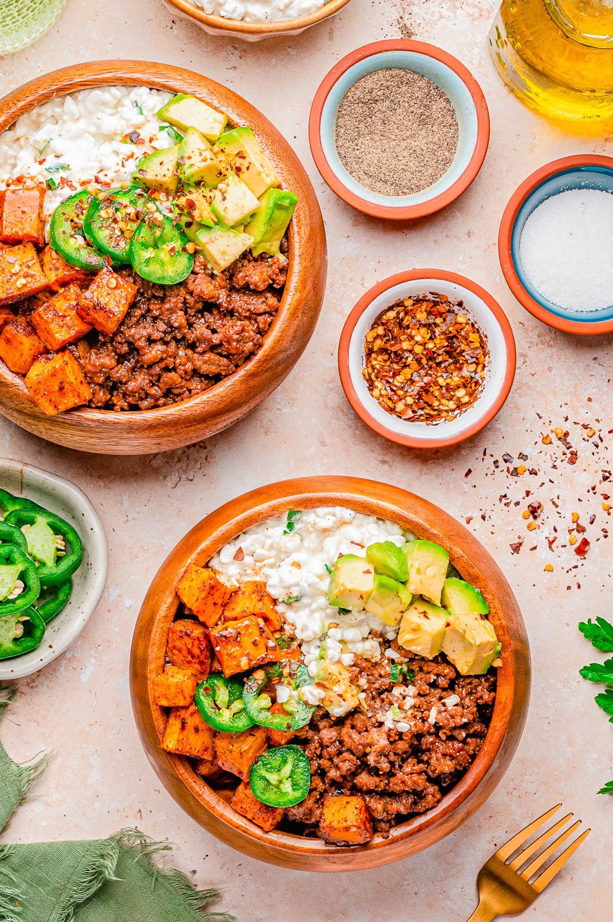 Two wooden bowls filled with ground beef, roasted sweet potatoes, diced avocado, jalapeño slices, and cottage cheese, surrounded by small bowls of seasonings and a gold fork.