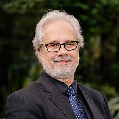 Homem de cabelos e barba brancas, usando óculos de aros pretos, vestindo terno e camisa pretas, com gravata azul, em frente a uma área de vegetação Homem de cabelos e barba brancas, usando óculos de aros pretos, vestindo terno e camisa pretas, com gravata azul, em frente a uma área de vegetação