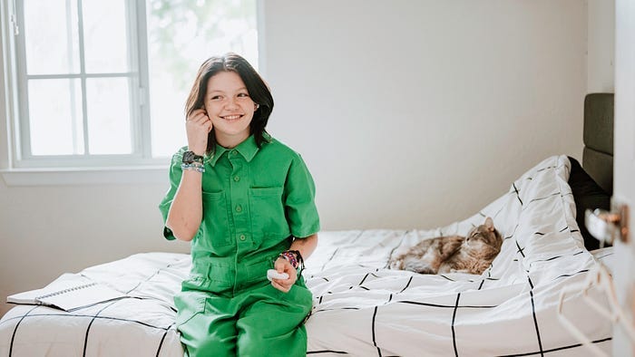 Nonbinary teen in a green jumpsuit, on her bed