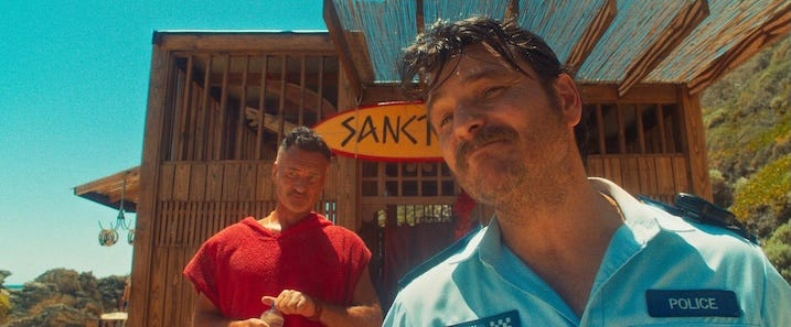 Police Officer & Surfer Clash Alt text: Scene from “The Surfer” where a local police officer smirks in the foreground while Julian McMahon stands behind him outside a wooden shack with a “Sanctuary” surfboard sign.