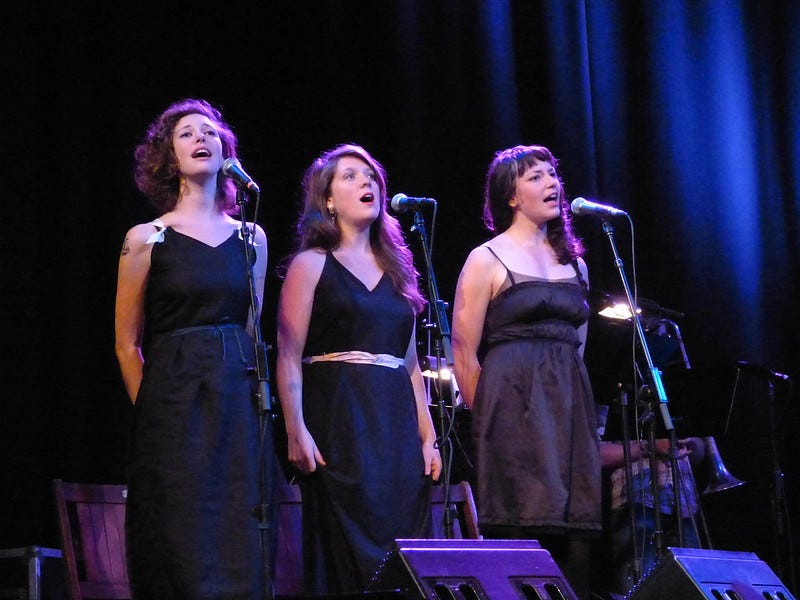 Three white women dressed in black as the fates sing into microphones https://www.flickr.com/photos/dan_tappan/6369771315/in/album-72157628066020835