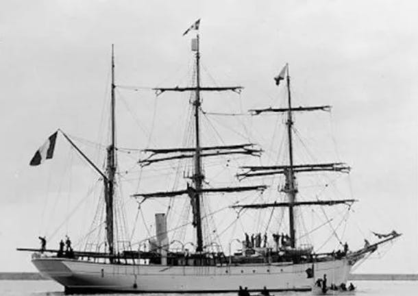 The 3-master sailing vessel Pourquoi Pas with its sails lowered, standing in shallow water. Sever members of the crew are on the  deck or in small boats below.