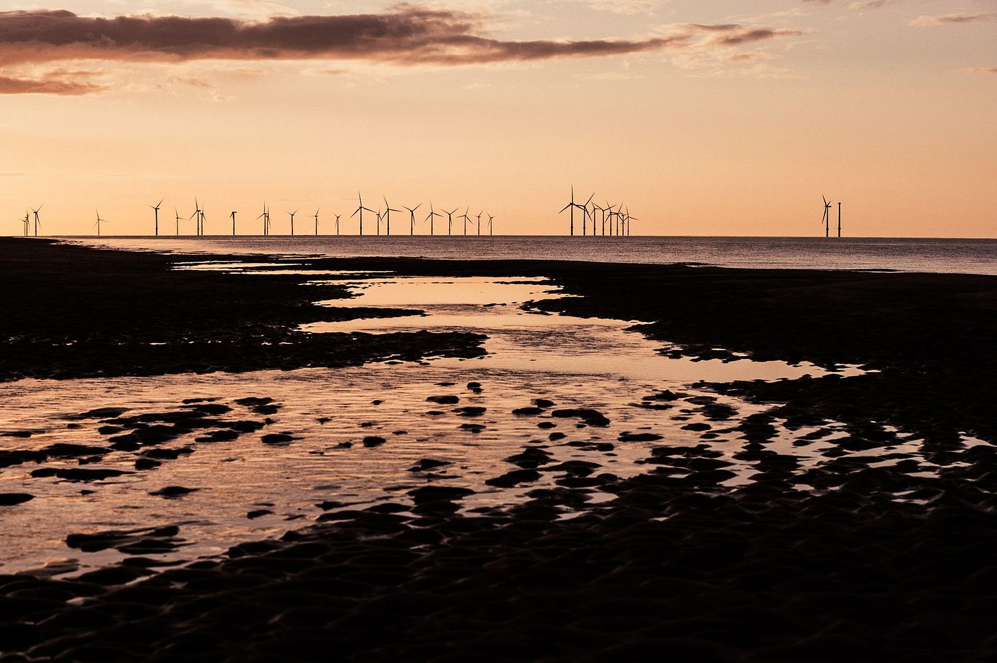 Wind turbines on the horizon with a river of water filtering through the sand below while the sun sets.