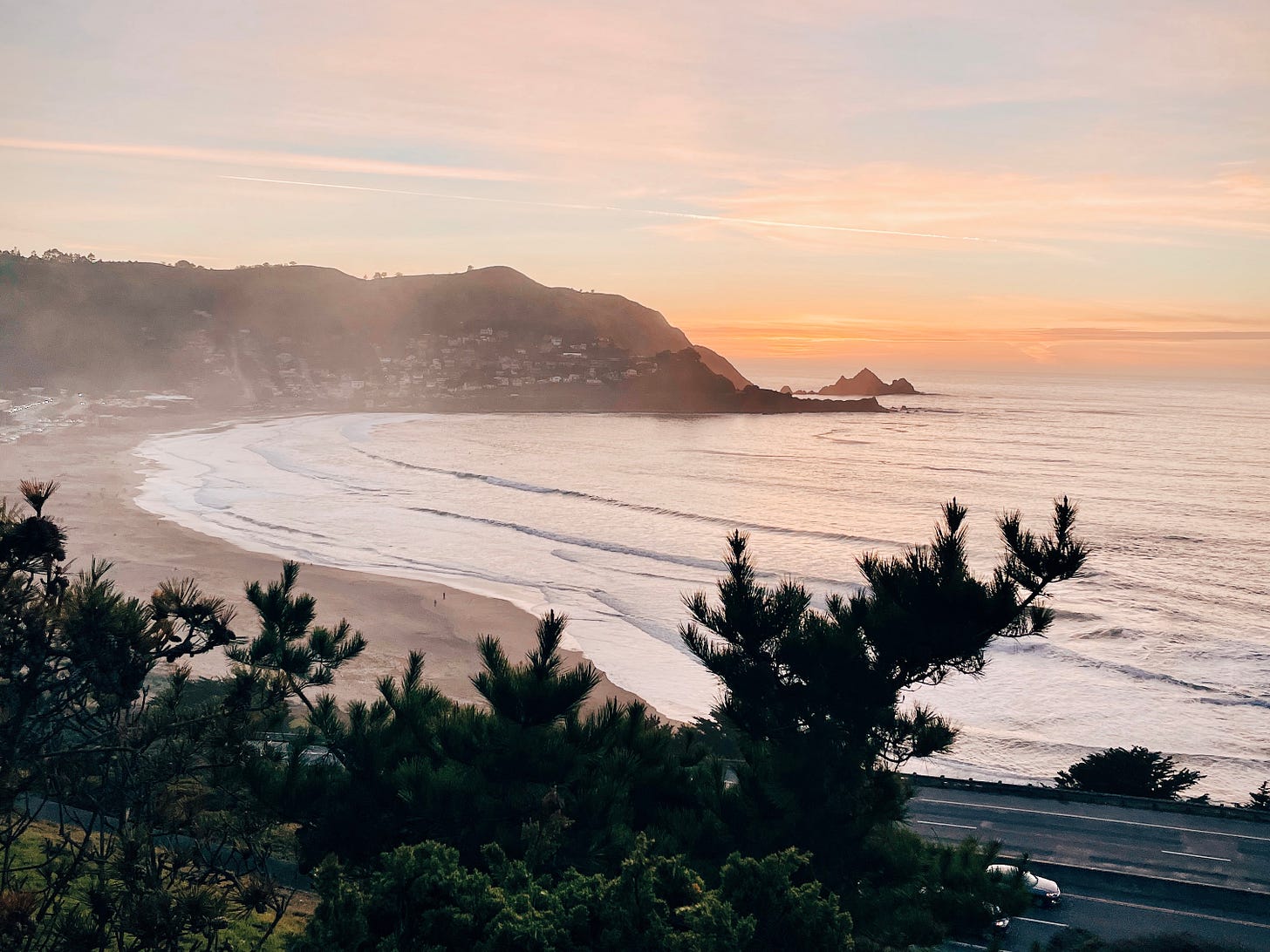 A photo: View from a cliff, looking onto a beach with a sunset and hills in the background.