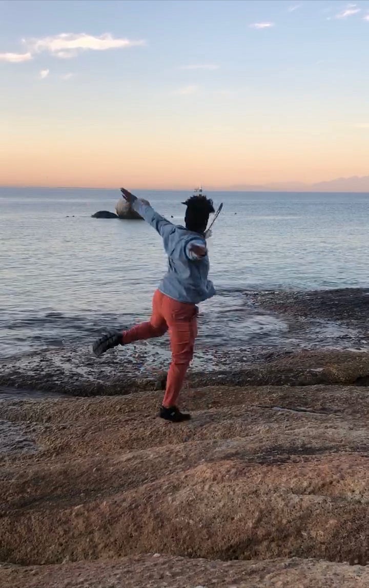 Two images of me moving across the ocean in Cape Town, South Africa. I'm wearing orange pants and a blue shirt. In the distance is an orange sky, the mountain line, boulders and faint blue water.