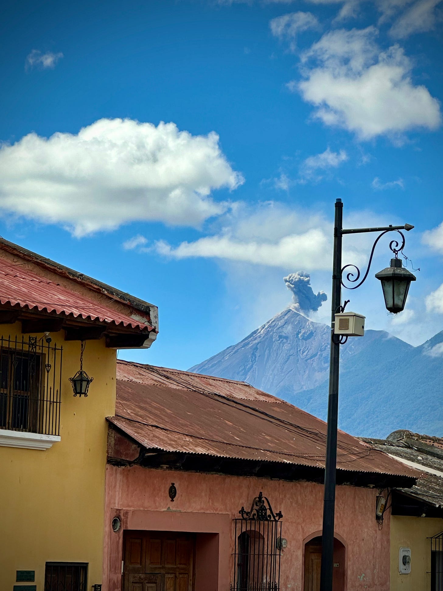 A lamppost with colorful buildings nearby and a volcano erupting ash and smoke in the distance.