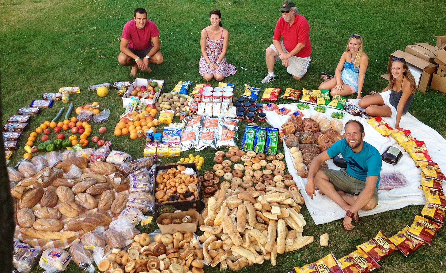 Six people sitting around a huge amount of food they've arranged in a circle on the lawn of a park.