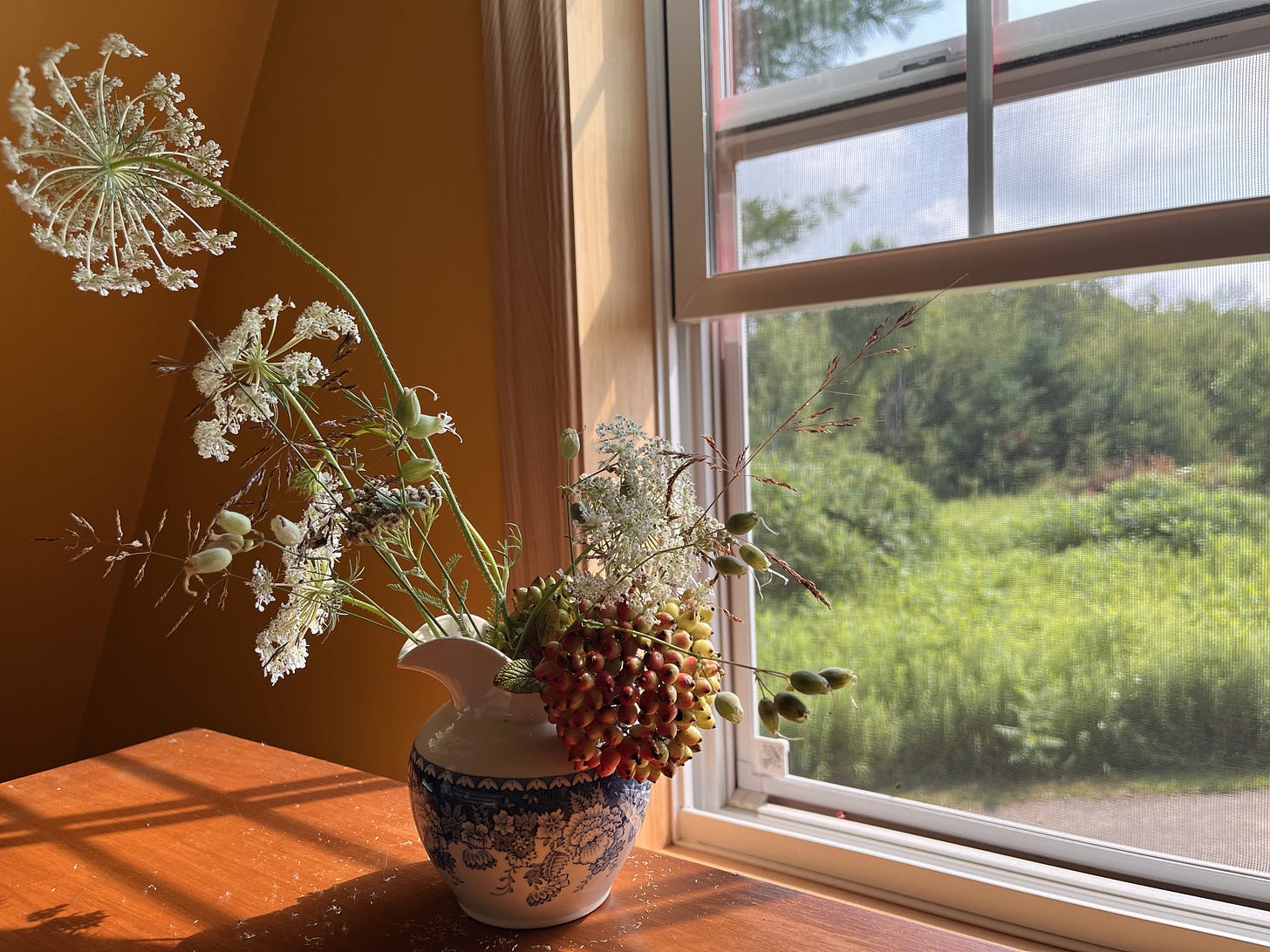 a small jug of wildflowers sits beside and open window looking onto fields