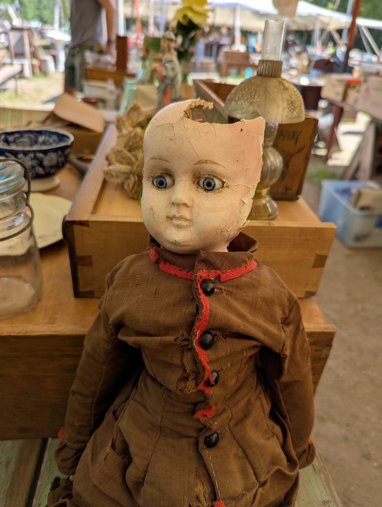 I might be too terrified to write this alt text. This is a photo of a doll at a flea market. The back of her head is broken off, jaggedly. She stares to the side, haunted, sad, bereft. She's wearing what I'm seeing now is an incredible brown canvas coat with red trim and buttons. I was too scared, previously, to notice the coat. I might be too terrified to write this alt text. This is a photo of a doll at a flea market. The back of her head is broken off, jaggedly. She stares to the side, haunted, sad, bereft. She's wearing what I'm seeing now is an incredible brown canvas coat with red trim and buttons. I was too scared, previously, to notice the coat.