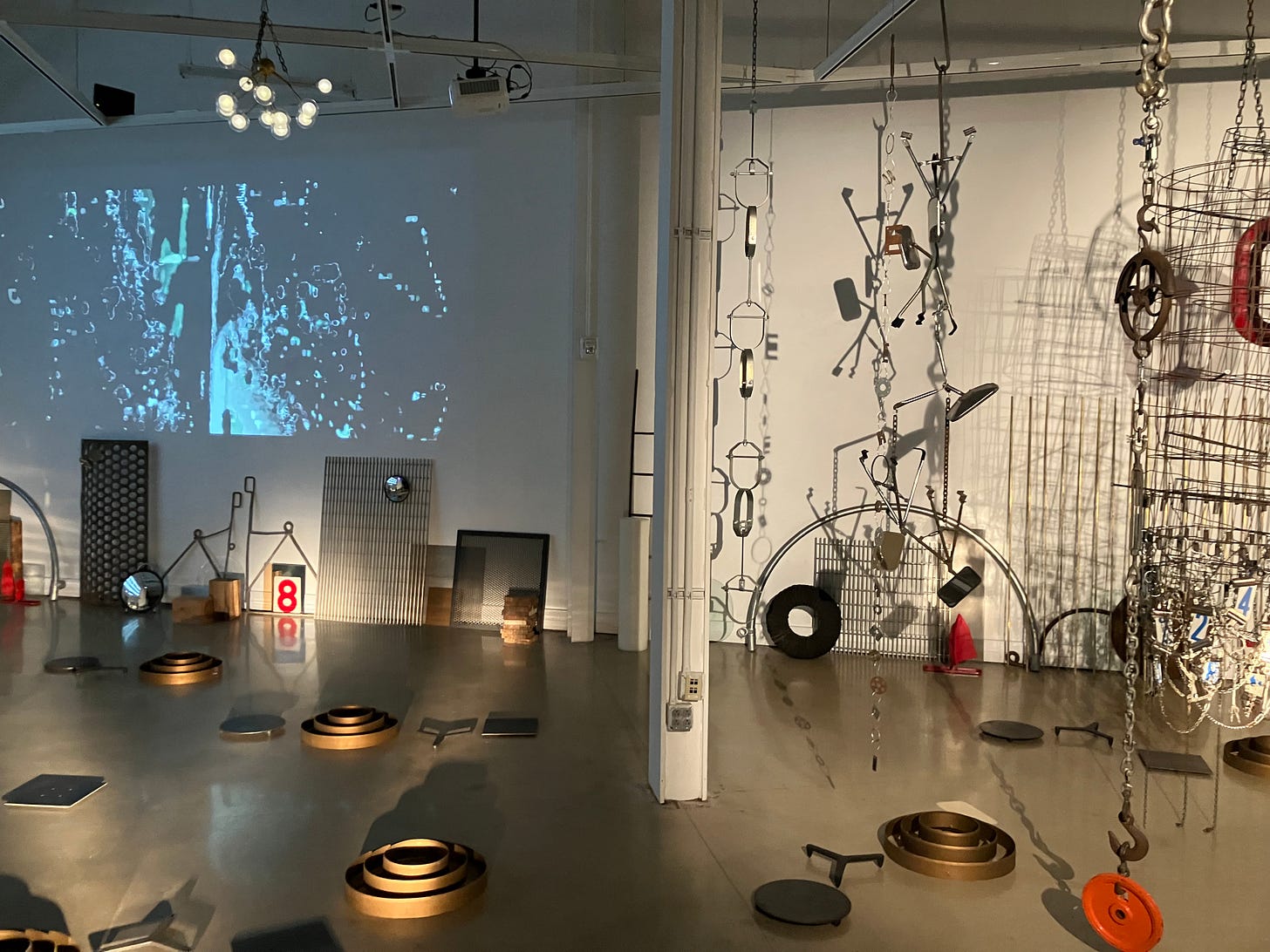 In a space that is half dark and half light, numerous objects, largely metal hardware, are scattered across the floor and leaning against the wall. On the left, an abstract, black and white video is projected upon the dark wall, while at right a group of vertical accumulations of metal objects hang from the ceiling.