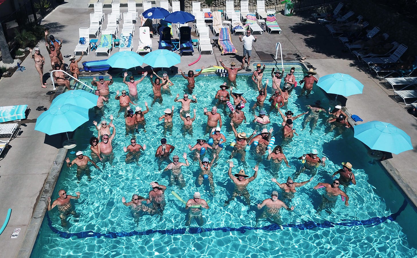 An aerial view of dozens of nude people gathered in a large outdoor swimming pool on a sunny day, most smiling and waving up at the camera. The pool is ringed with turquoise umbrellas, white lounge chairs, and towels at a sun club resort. An aerial view of dozens of nude people gathered in a large outdoor swimming pool on a sunny day, most smiling and waving up at the camera. The pool is ringed with turquoise umbrellas, white lounge chairs, and towels at a sun club resort.