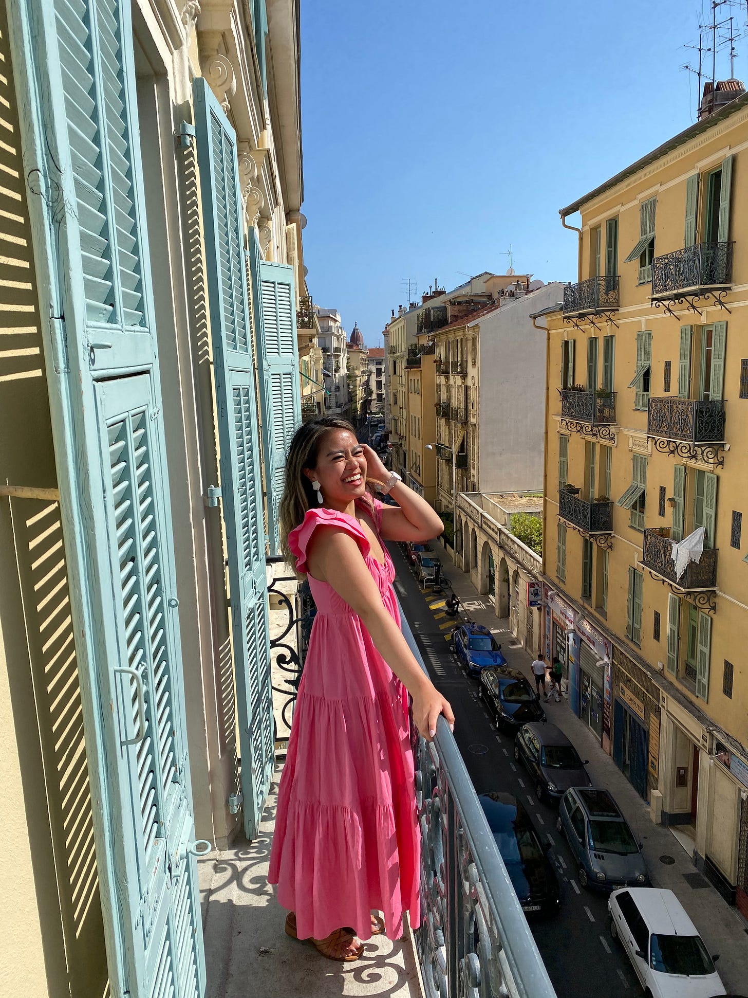 Joelle wearing a bright pink maxi dress smiling and standing on a light blue and white skinny balcony overlooking a street in Nice, France