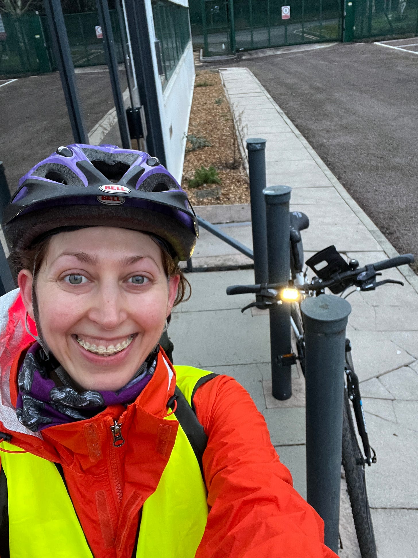 A photo of me wearing my helmet and high vis jacket proudly smiling stood next to my bike outside of the office.