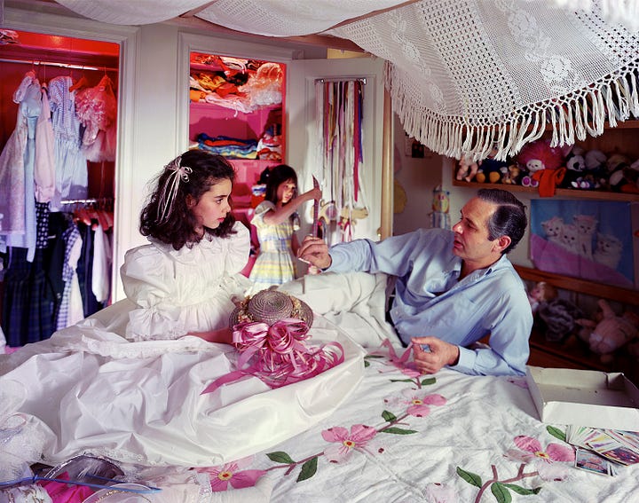 1: Photograph by Tina Barney depicting a bed covered in a white embroidered bedspread adorned with pink flowers. A young girl in a white dress with puffed sleeves and her hair adorned with ribbons sits on the bed. Next to her is a man in a blue shirt, leaning on the bed, appearing to engage in conversation with her. In the background, another girl in a pastel-colored dress stands in a brightly lit closet filled with clothes hanging on a rod. 2: Photograph by Tina Barney depicting two women in a room with cream colored walls. On the left, one woman with wavy brown hair is seated, turned toward the camera, resting her chin on her hand. To the right, facing a mirror, a young woman with short, bleach blonde hair applies bright red lipstick. She is also dressed in black and stands in a profile position, reflecting in the mirror. 3: Photograph by Tina Barney, depicting three people engaged in conversation in a sunlit room. On the left, an older man with white hair wearing a black vest over a white shirt gestures towards the center. In the middle, a young person stands with their hands holding the back of their neck. To the right, a woman in a light brown blouse and white skirt holds her chin thoughtfully. 4: Photograph by Tina Barney featuring two women in a pastel pink bathroom, both wearing pink robes. On the left, one woman with short dark hair and glasses gazes toward the camera, her hand on her hip. She is standing in front of a mirror, in which her reflection is visible. To the right, a young woman with long dark hair peeks from behind floral curtains covering a window.