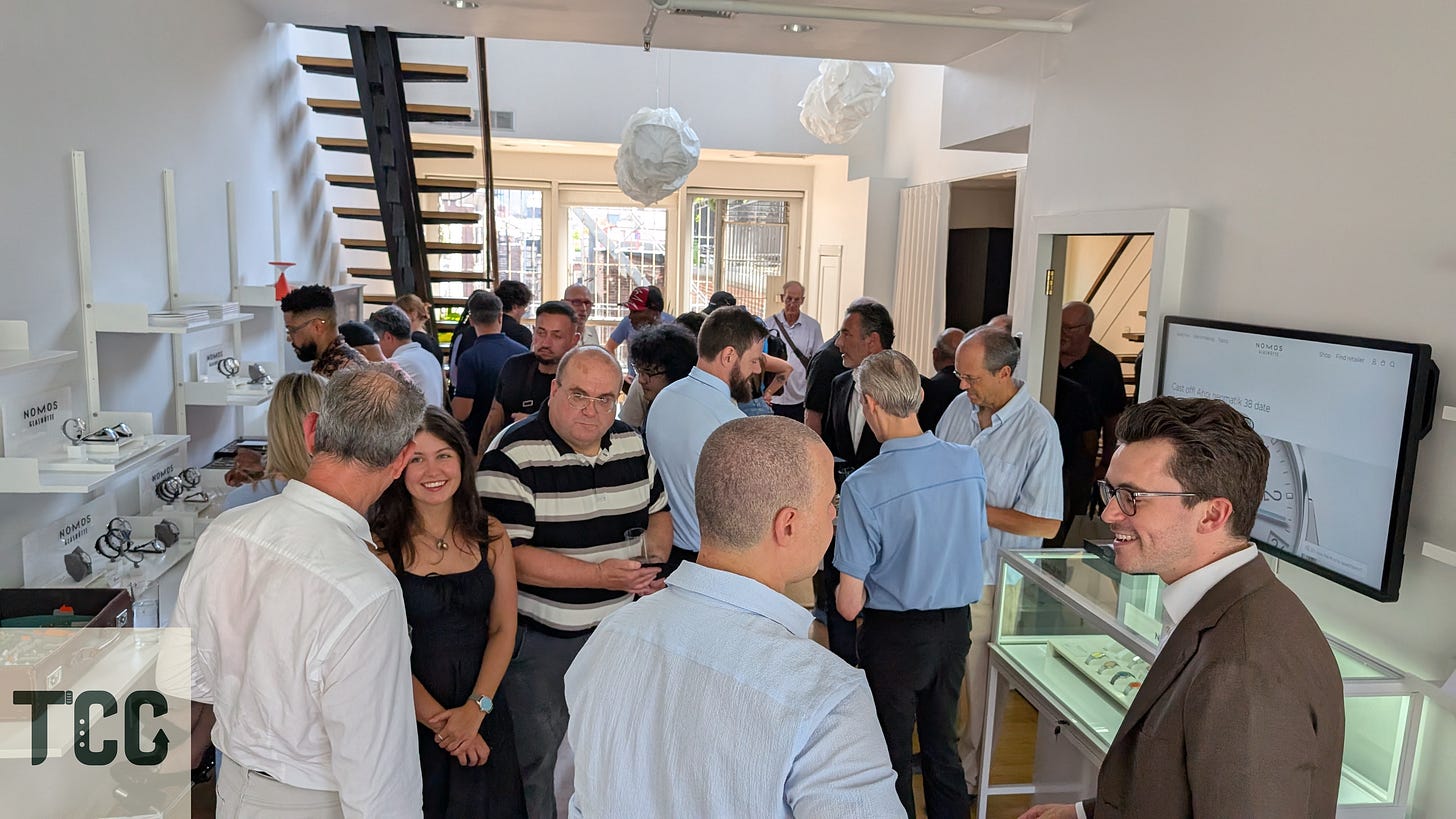 A large group of people inside the NOMOS Lounge in New York City, socializing and examining watches displayed on wall shelves. A staircase leads to a loft, and natural light fills the space through rear windows.