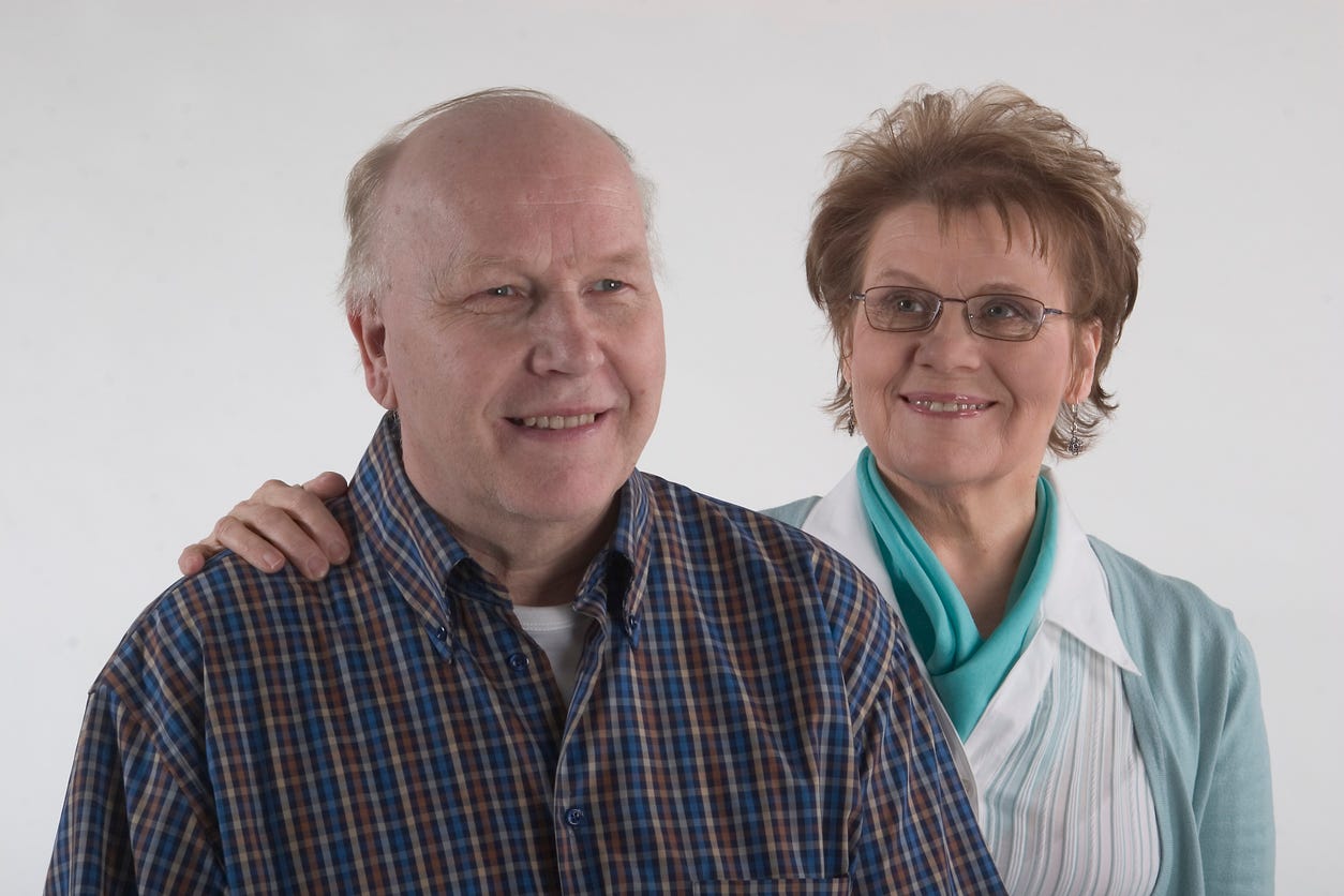 Smiling, not-too-ederly couple, she with her hand on his right shoulder. He's wearing plaid shirt. Her outfit is blue-green.