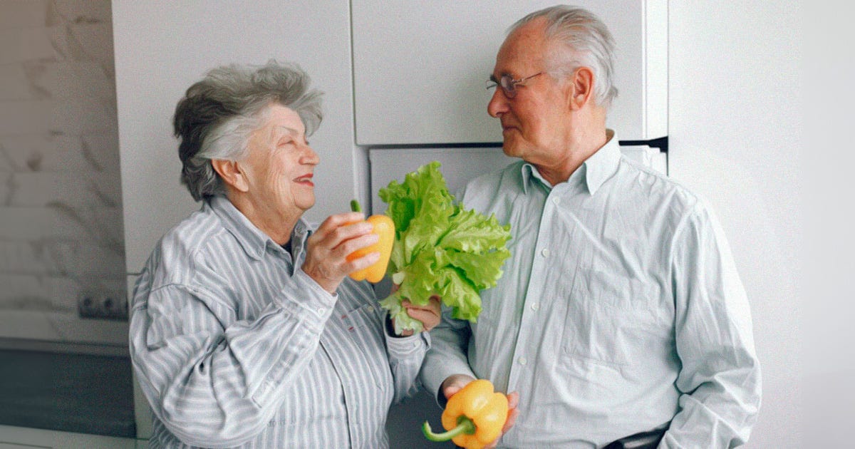 Imagem de um casal de idosos conversando. A mulher segura nas mãos um pimentão e um pé de alface, enquanto o homem também segura um pimentão com a mão direita. Ambos vestem roupas claras