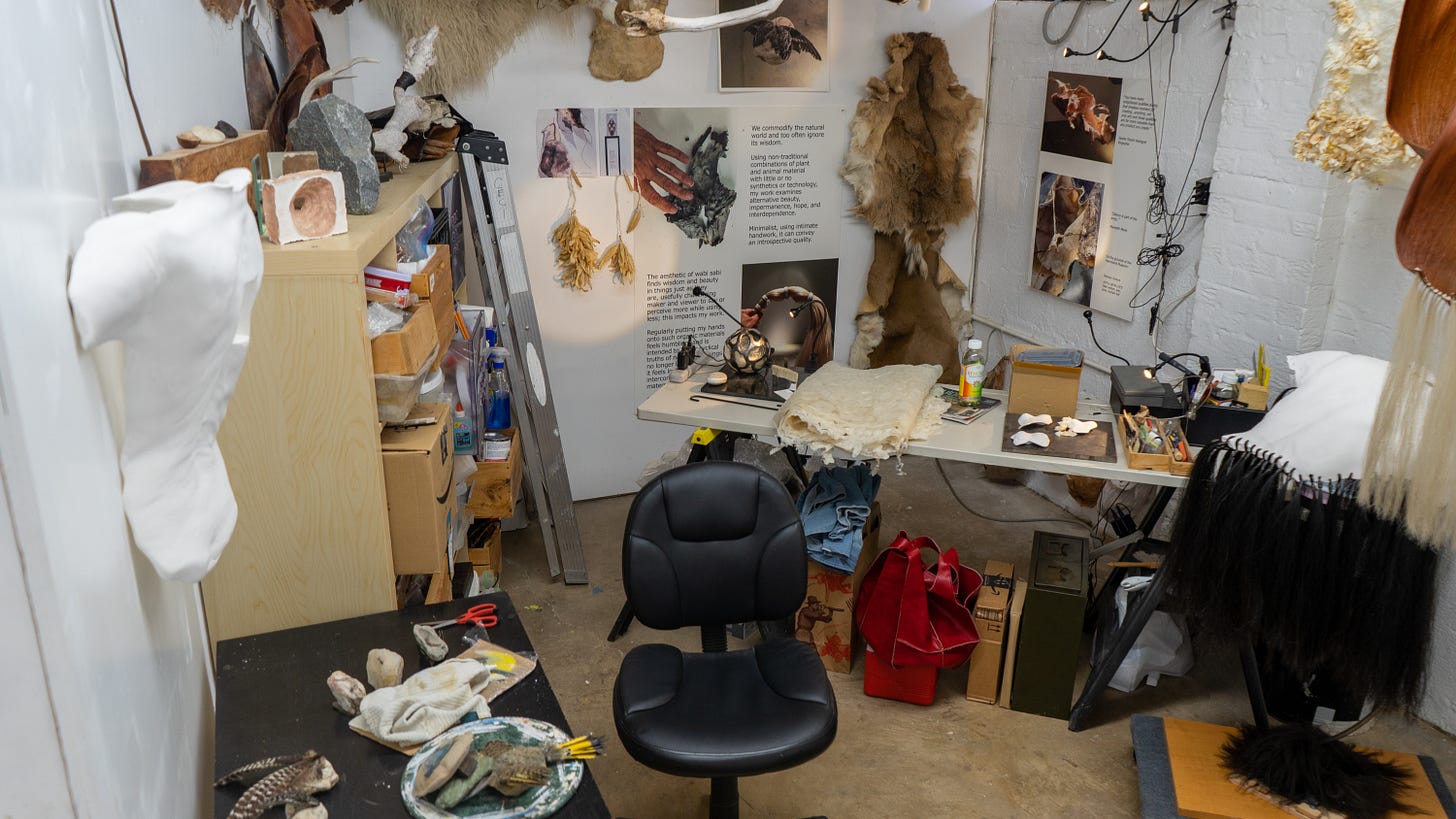 In Ceci Cole McInturff’s studio, there are two desks, one on the left against the wall, and another in the corner across from the entrance. The desks carry scattered materials - feathers, rocks, homemade paper, notes, pens, and small tool boxes. On the walls are works in various stages of creation, some complete. There is a hide hanging on the back wall, posters with text from previous shows, and dried organic material. On the left side is a cast of an upper back in stark white. Bookshelves hold boxes and boxes, bursting. Cole McInturff’s empty black office chair sits open at the front center of the image.