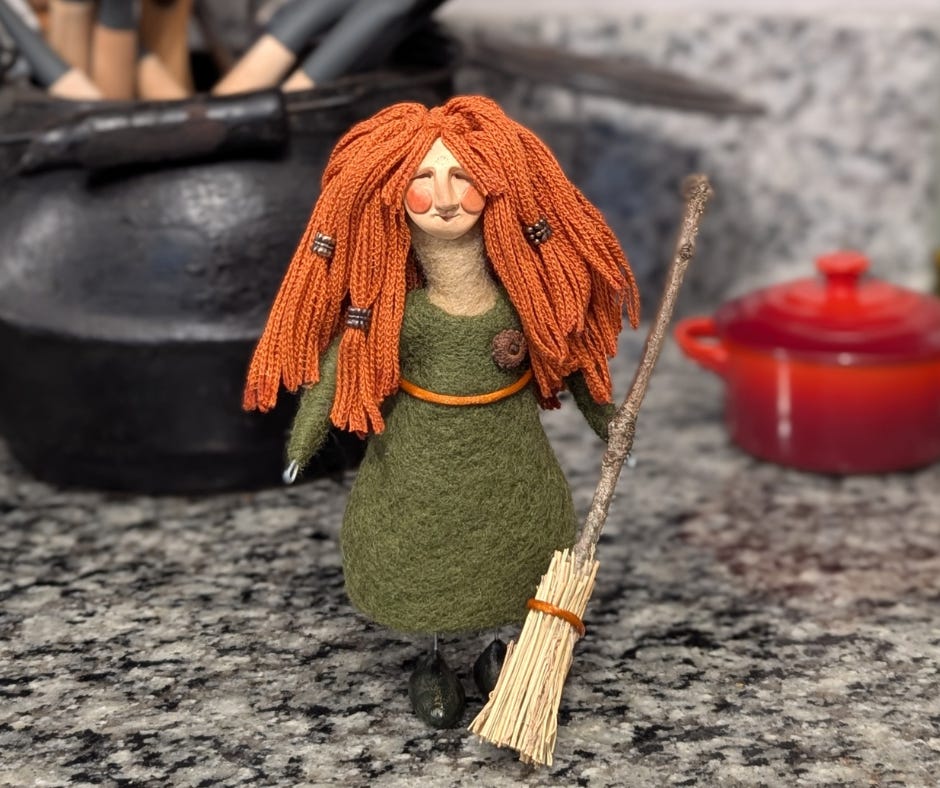 A needle felted kicthen witch doll with a green dress and orange haor holding a broom standing on a kicthen counter. A quirky doll made by Stacy Vajta