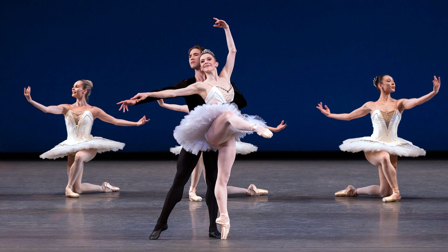 Megan Fairchild, in a white tutu and tiara, and Joseph Gordon, in black tights and tunic, perform a ballet pas de deux onstage. Three women in white tutus are behind them kneeling with their arms open. Fairchild does an attitude devant on pointe and rests her right hand on Gordon's extended right arm as he stands behind her in tendu, holding her around the waist with his left arm. Megan Fairchild, in a white tutu and tiara, and Joseph Gordon, in black tights and tunic, perform a ballet pas de deux onstage. Three women in white tutus are behind them kneeling with their arms open. Fairchild does an attitude devant on pointe and rests her right hand on Gordon's extended right arm as he stands behind her in tendu, holding her around the waist with his left arm.