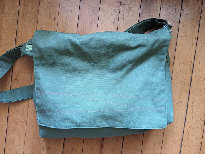 Five photos of the Mythical Messenger Bag. In the first, I (a white lady, aged 52) am standing in the hallway wearing the bag. The other photos are of the bag on its own. The bag is olive green canvas with an adjustable strap. There is hot pink decorative stitching. There is an exterior welt pocket on the back with a hot pink zipper. There is a pocket on the front with a blue square patch that says "Make Art" in a red cursive font. The inner lining fabric is floral -- blue, green, coral line drawn flowers on a white background. Inside the bag, there are two pockets. One is a zippered pocket with a hot pink zipper. The other is a divided pocket with slots for pens, a phone, and something else roughly cell-phone-sized.