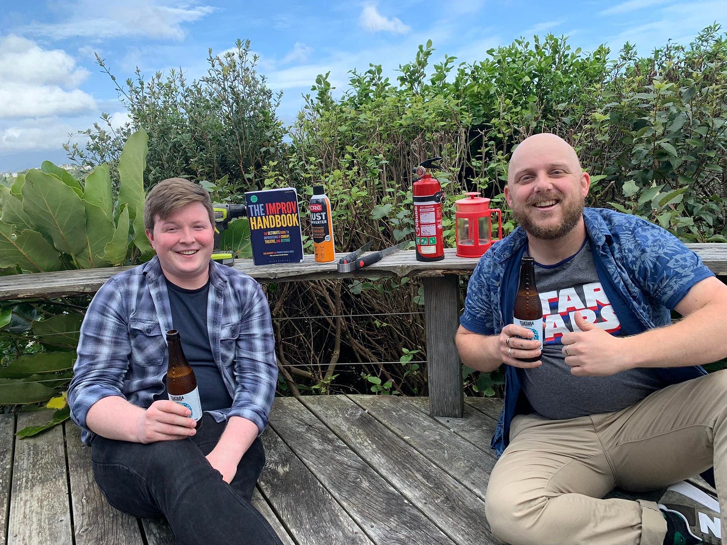 Austin and Dylan, two young white men, sit on a deck with beers and camping equipment. THere is a copy of The Improv Handbook on the deck with them. 