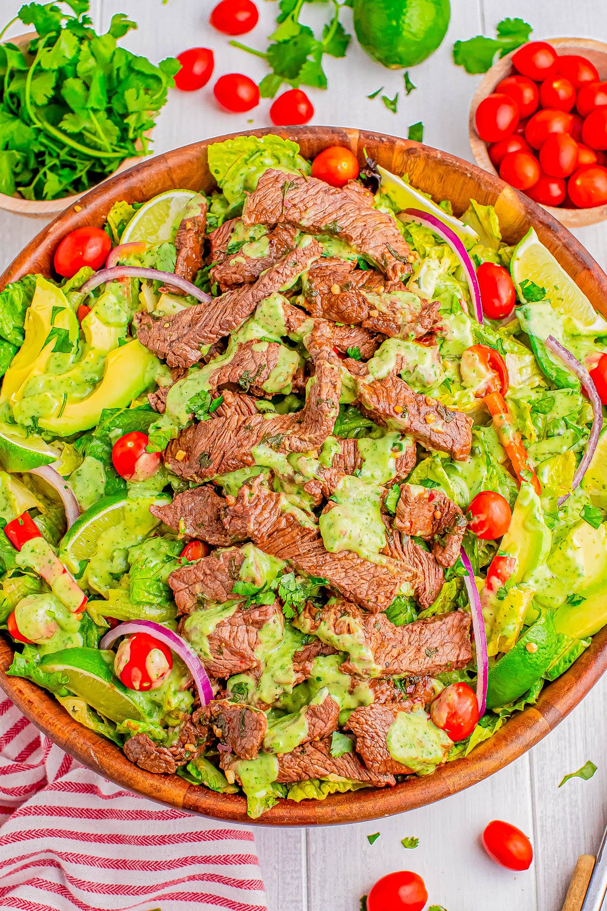 A wooden bowl filled with a salad featuring sliced steak, cherry tomatoes, avocado, red onion, and a green dressing. Other ingredients like parsley and lime are visible in the background. A wooden bowl filled with a salad featuring sliced steak, cherry tomatoes, avocado, red onion, and a green dressing. Other ingredients like parsley and lime are visible in the background.